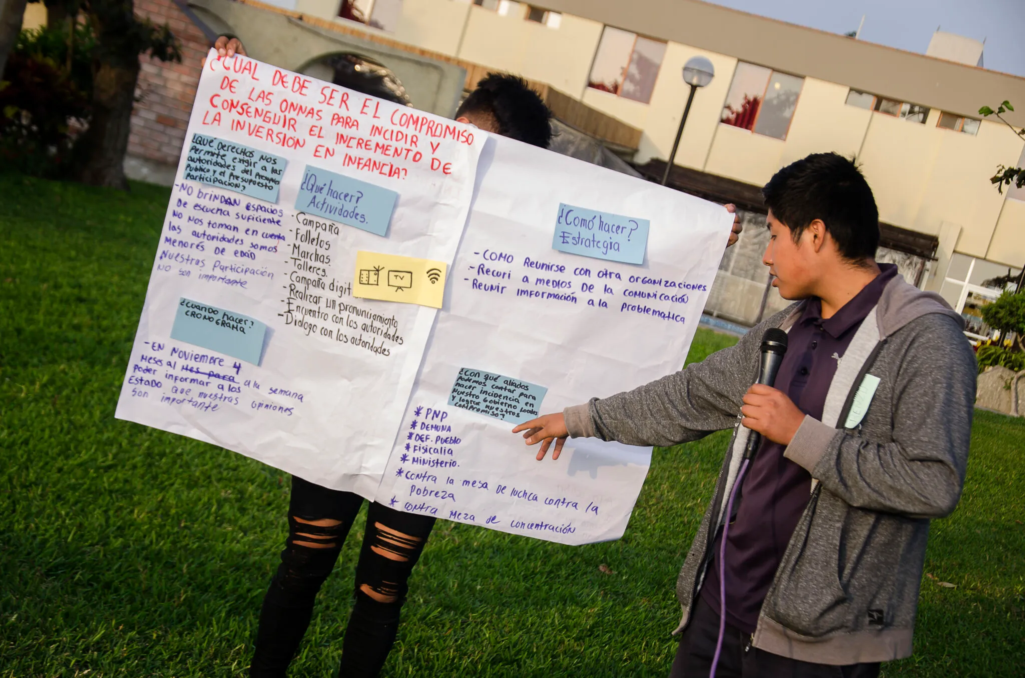 Dos adolescentes exponen ante papelógrafos al aire libre; uno habla por un micrófono.
