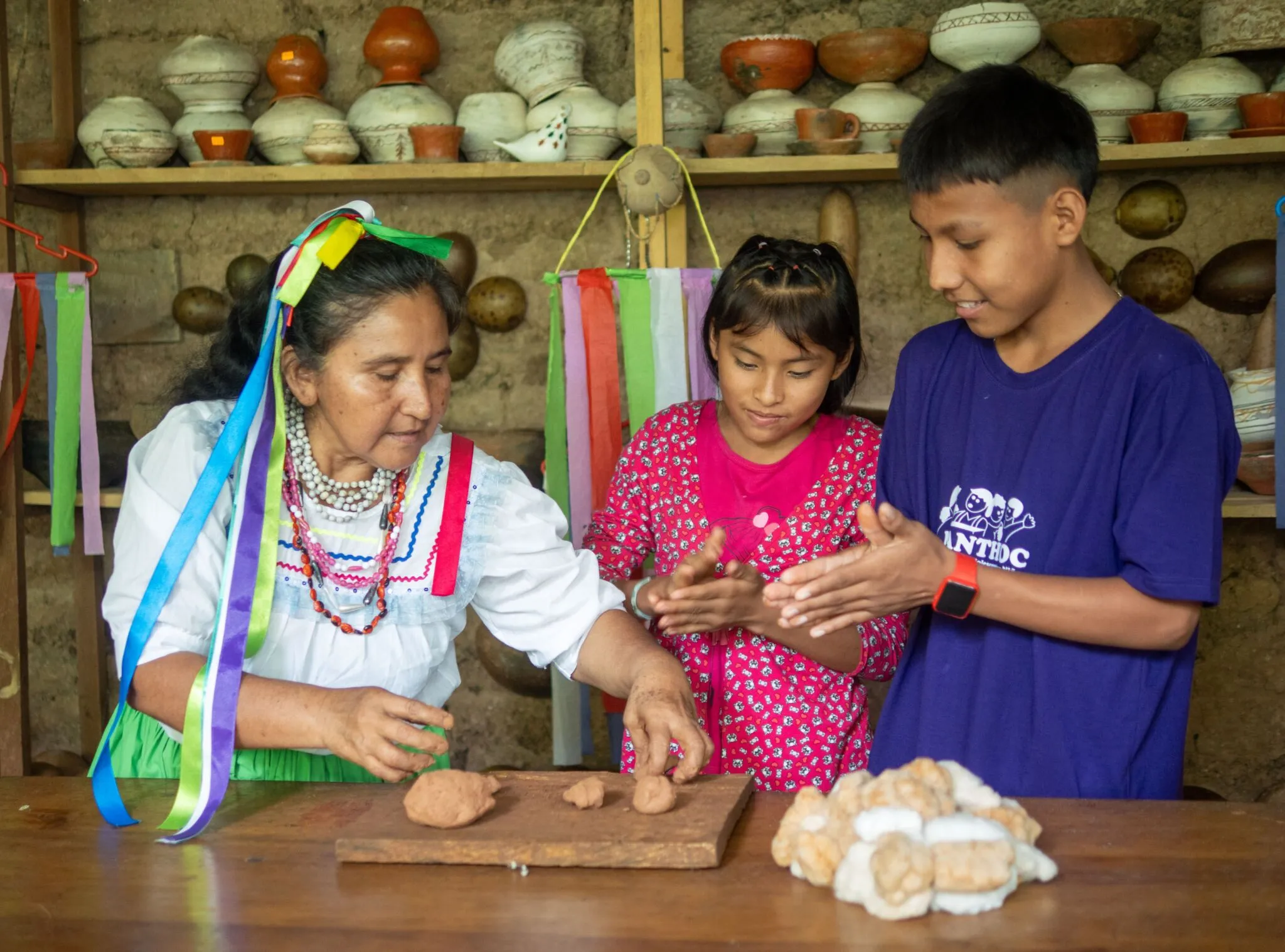 Una mujer mayor enseña a una niña y un niño a trabajar con las manos en una actividad intercultural.