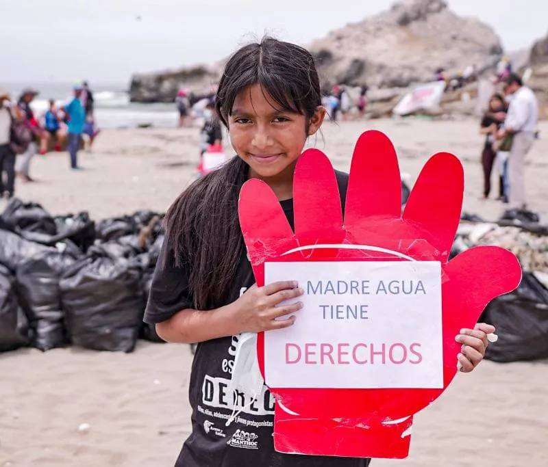 Una niña sonríe en la playa sosteniendo un cartel en forma de mano roja que dice «Madre agua tiene derechos».